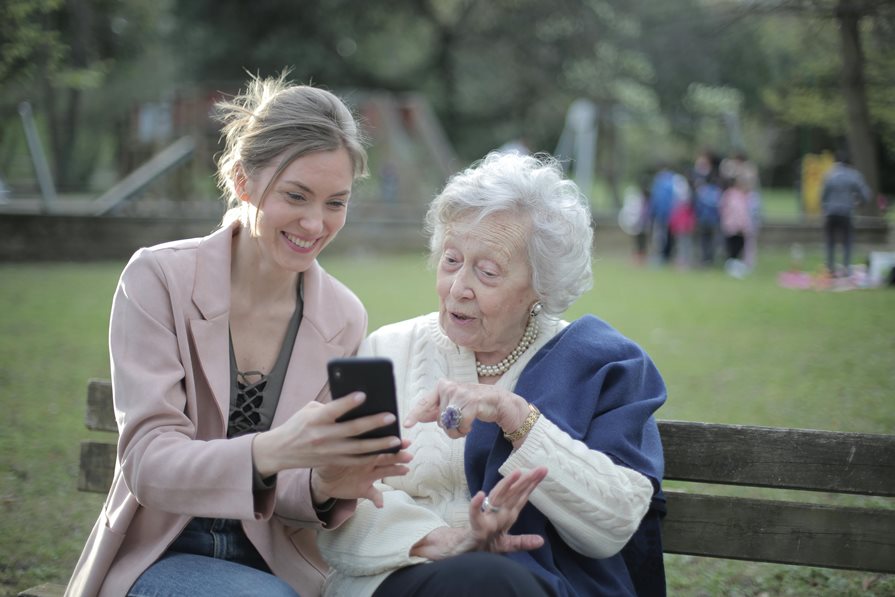 A younger person and a senior look at a phone on a park bench