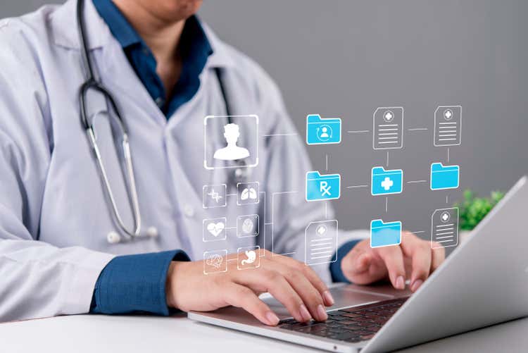 A health care provider types on a laptop while a graphic of a digital system floats above it.